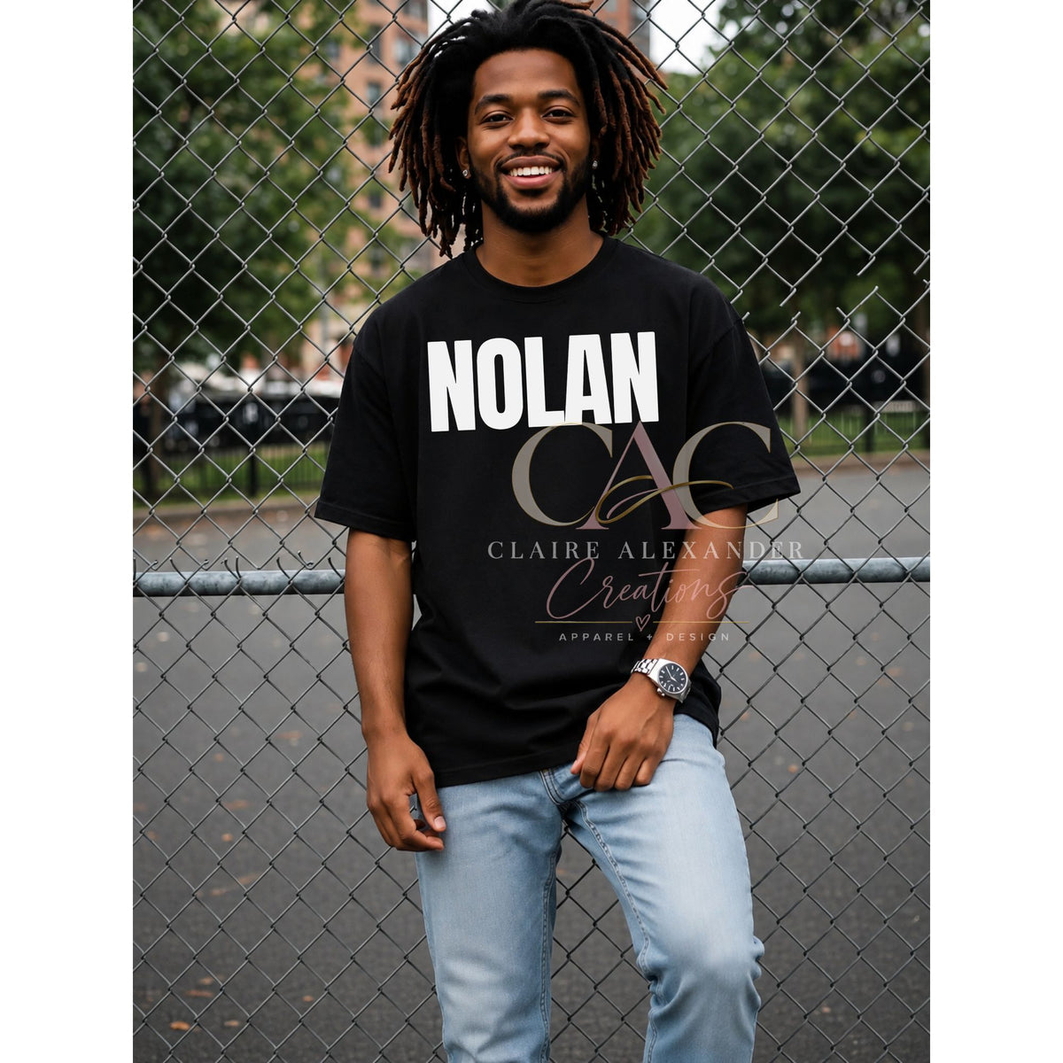 A man with dreadlocks stands in front of a chain-link fence wearing a black graphic t-shirt with 'NOLAN' and 'CLAIRE ALEXANDER CREATIVES APPAREL LTD' printed on the front, paired with light blue jeans.